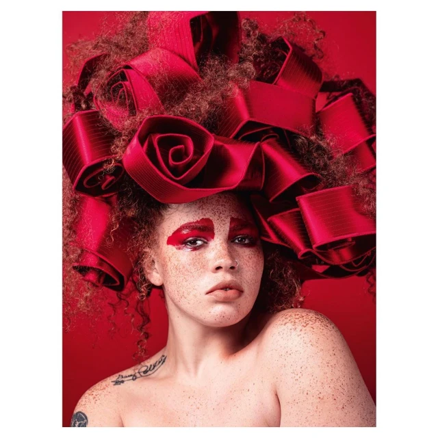 Close-up portrait of a woman with auburn hair and freckles, wearing a red ribbon hairstyle, on a matching vibrant red background.