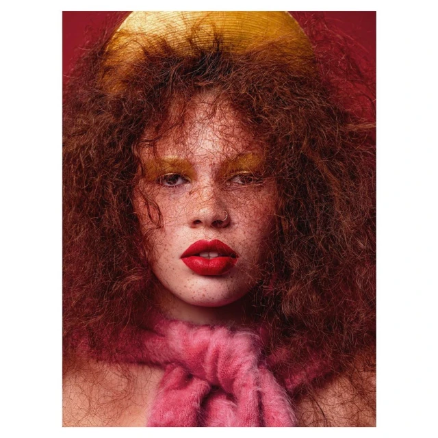 Portrait of an adult female model with red hair, dramatic makeup, and a gold hat posing in front of a red background in a photo studio.