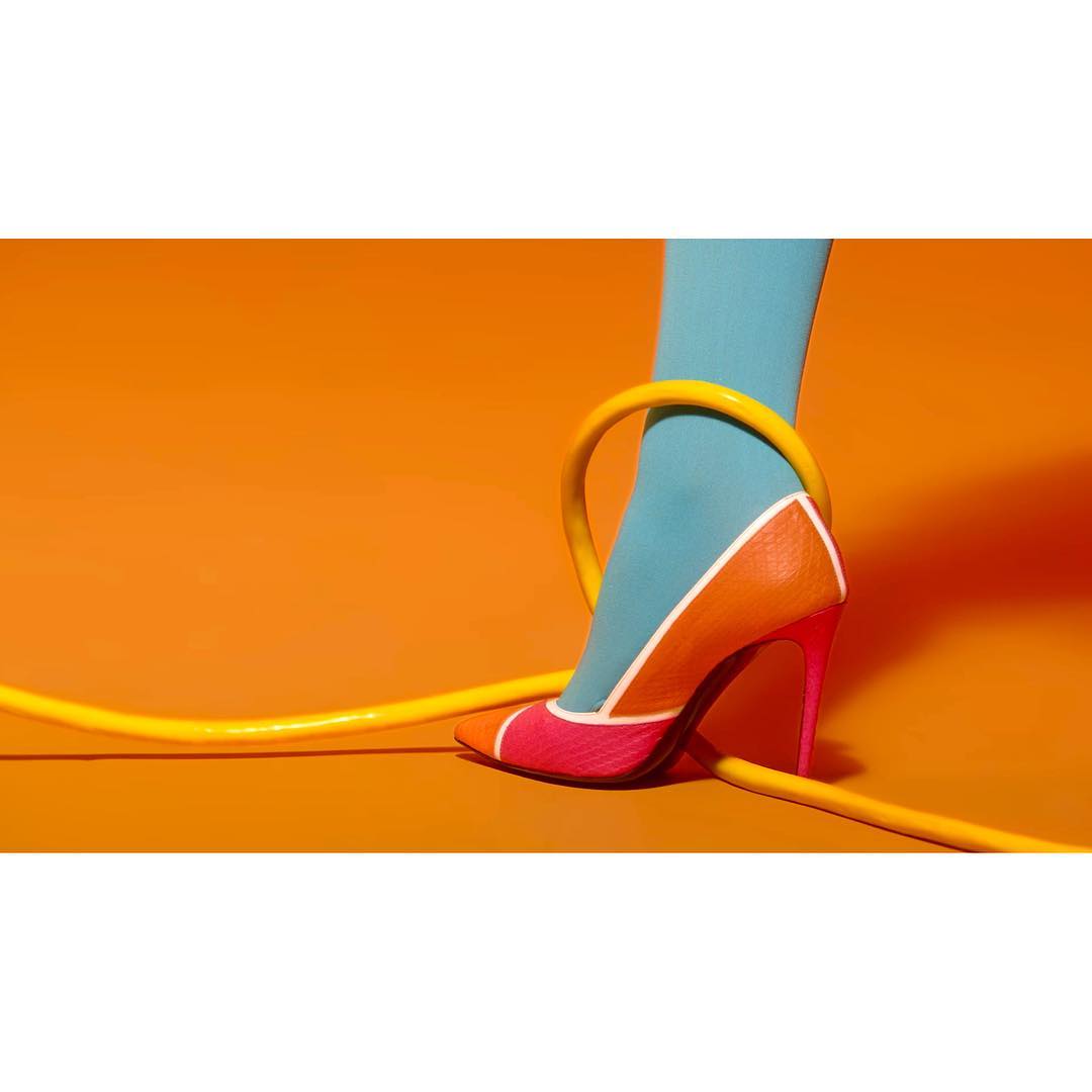 High heel pump shoe with orange and pink color block design, worn with blue stocking, and encircled by yellow tube, on orange background.