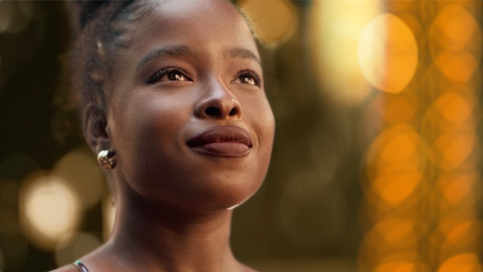 Close-up shot of a smiling Black woman with gold hoop earrings, against a blurred bokeh light background with a shallow depth of field.