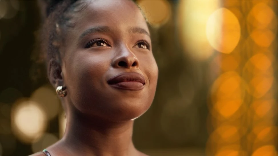 Close-up shot of a smiling Black woman with gold hoop earrings, against a blurred bokeh light background with a shallow depth of field.