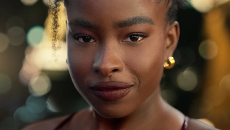 Close-up portrait of a Black woman with dark hair, minimal makeup, and small gold hoop earrings against a dark blurred background.