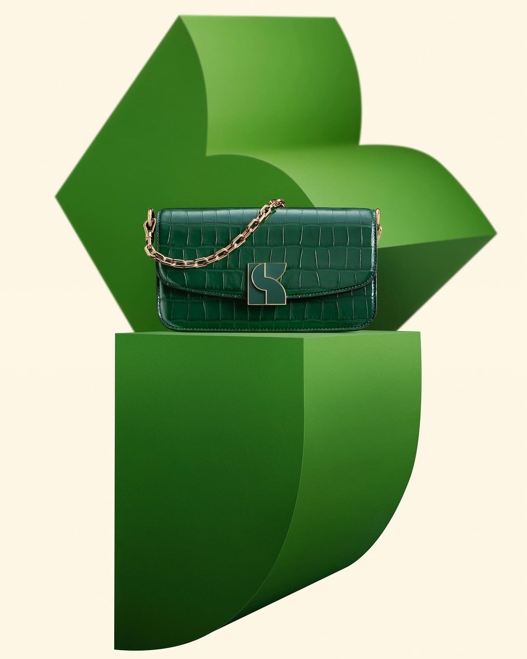 Elegant green crocodile-texture handbag with gold chain handle displayed on geometric green stands against a cream background.