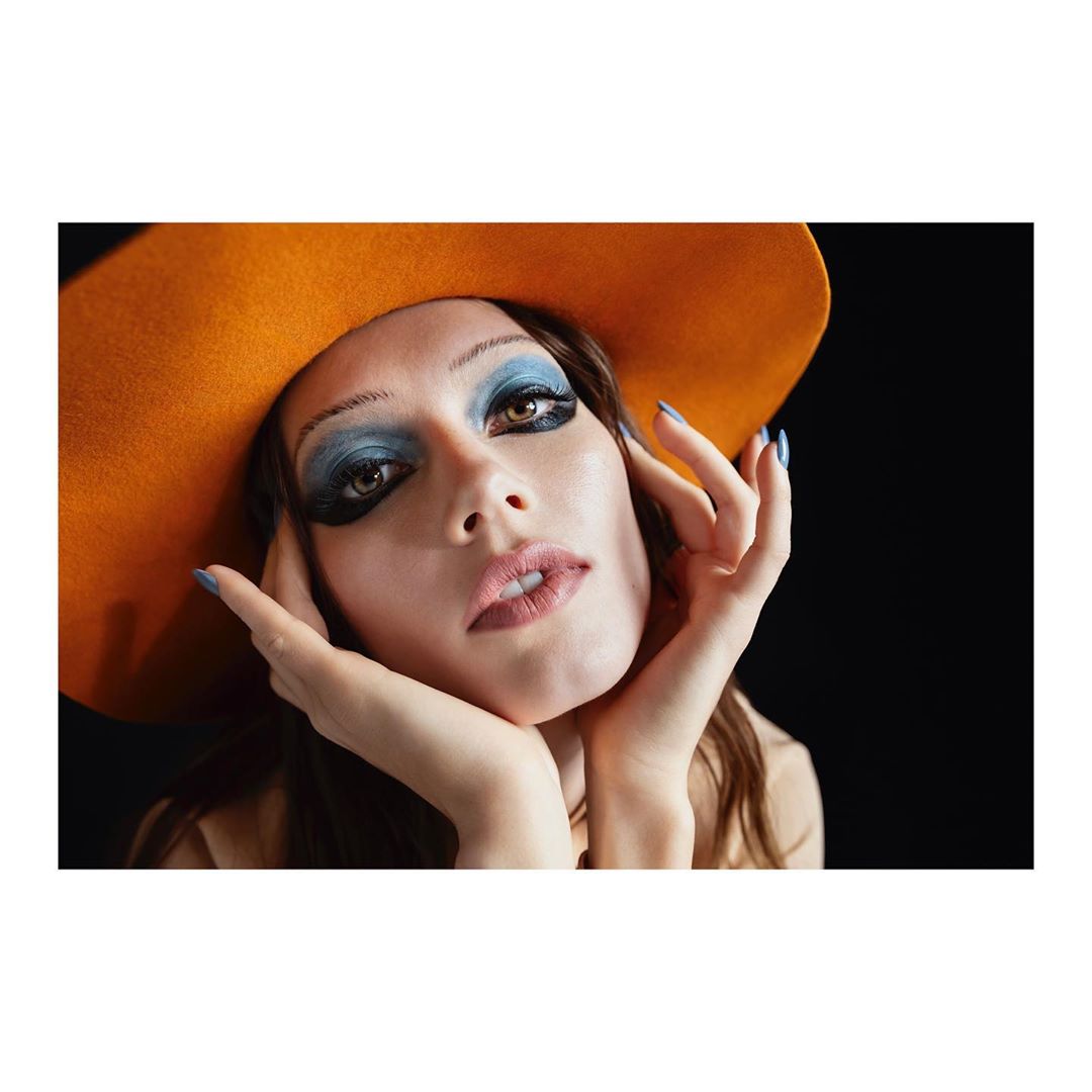 A woman with blue eyeshadow and an orange hat looks into the camera with hands framing her face in a studio setting on black background.