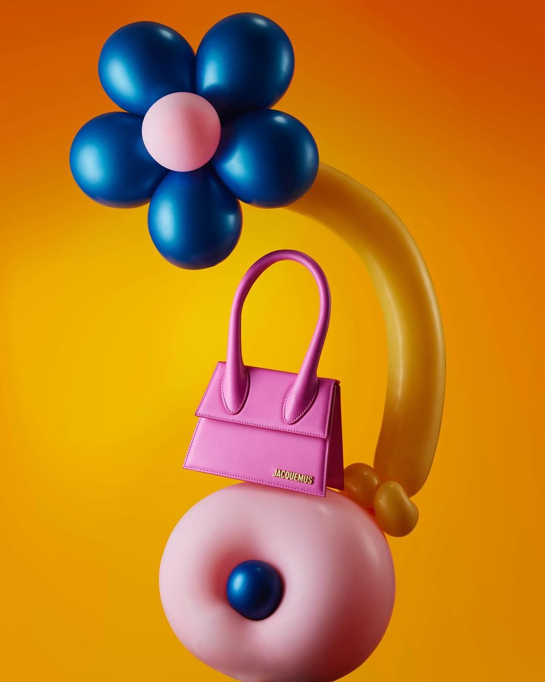 Pink Jacquemus purse atop a donut-shaped balloon with a blue-and-pink flower balloon arrangement, set against a vibrant yellow background.
