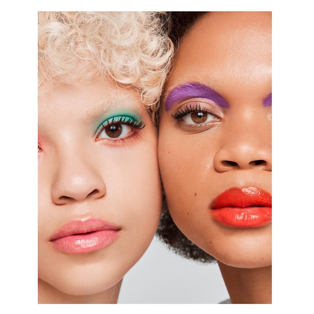 Close-up of two women with colorful makeup. One woman has green eyeshadow and pale skin; the other has purple eyeshadow and red lipstick.