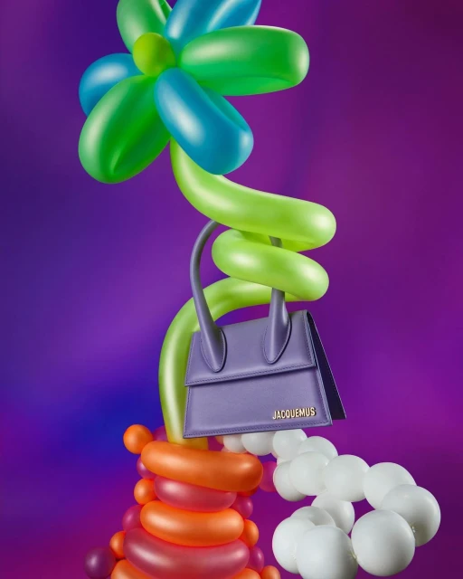 Fashion handbag displayed on a balloon flower sculpture against a gradient purple background in a studio product shot for accessories.