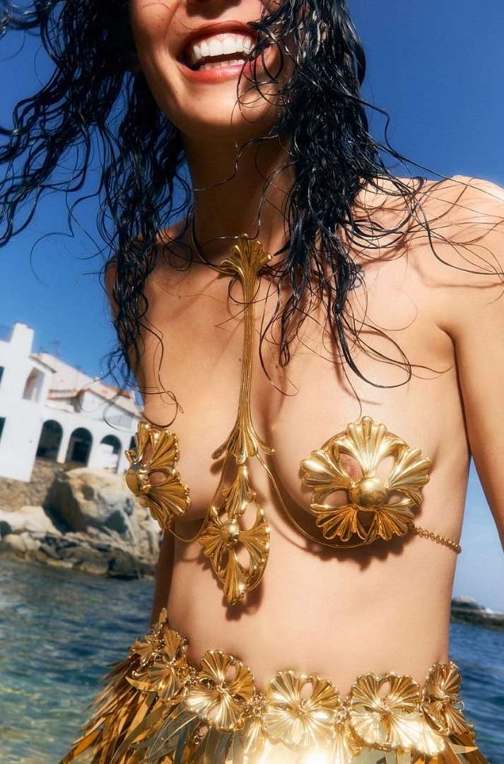 A woman smiles in golden, ornate swimwear with water in the foreground and a white building and blue sky in the background.