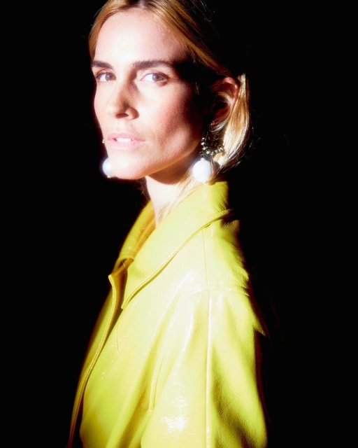 Woman in yellow jacket stands in a dark setting with bright light on her face and jacket, accessorized with dangling pearl earrings.