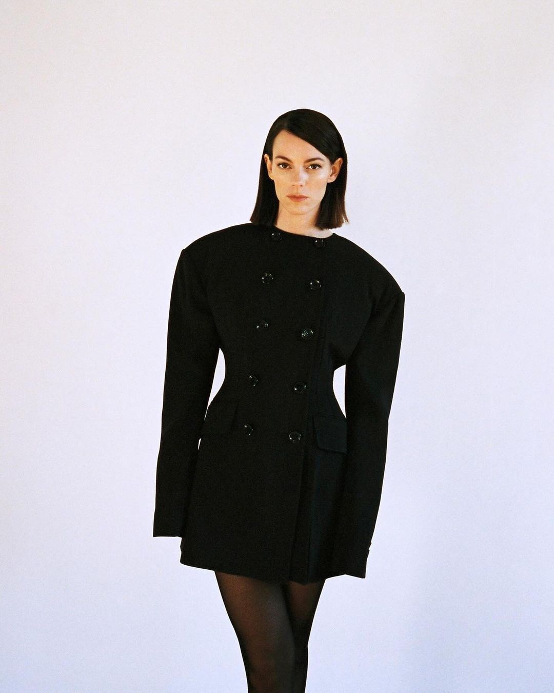 Brunette woman poses in a fitted black jacket dress against a white background, showcasing fashion and editorial style.