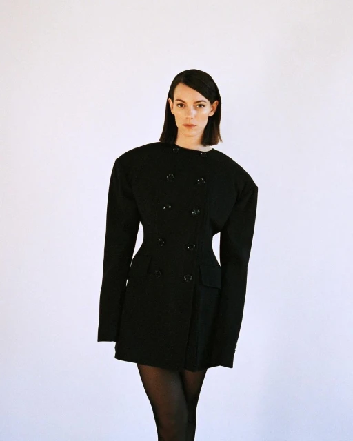 Brunette woman poses in a fitted black jacket dress against a white background, showcasing fashion and editorial style.