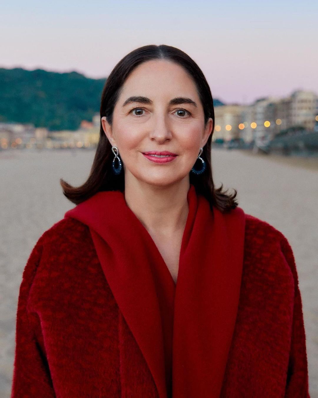 Portrait of an adult woman with dark hair wearing a red coat, posing on a sandy beach with an urban city skyline at dusk.