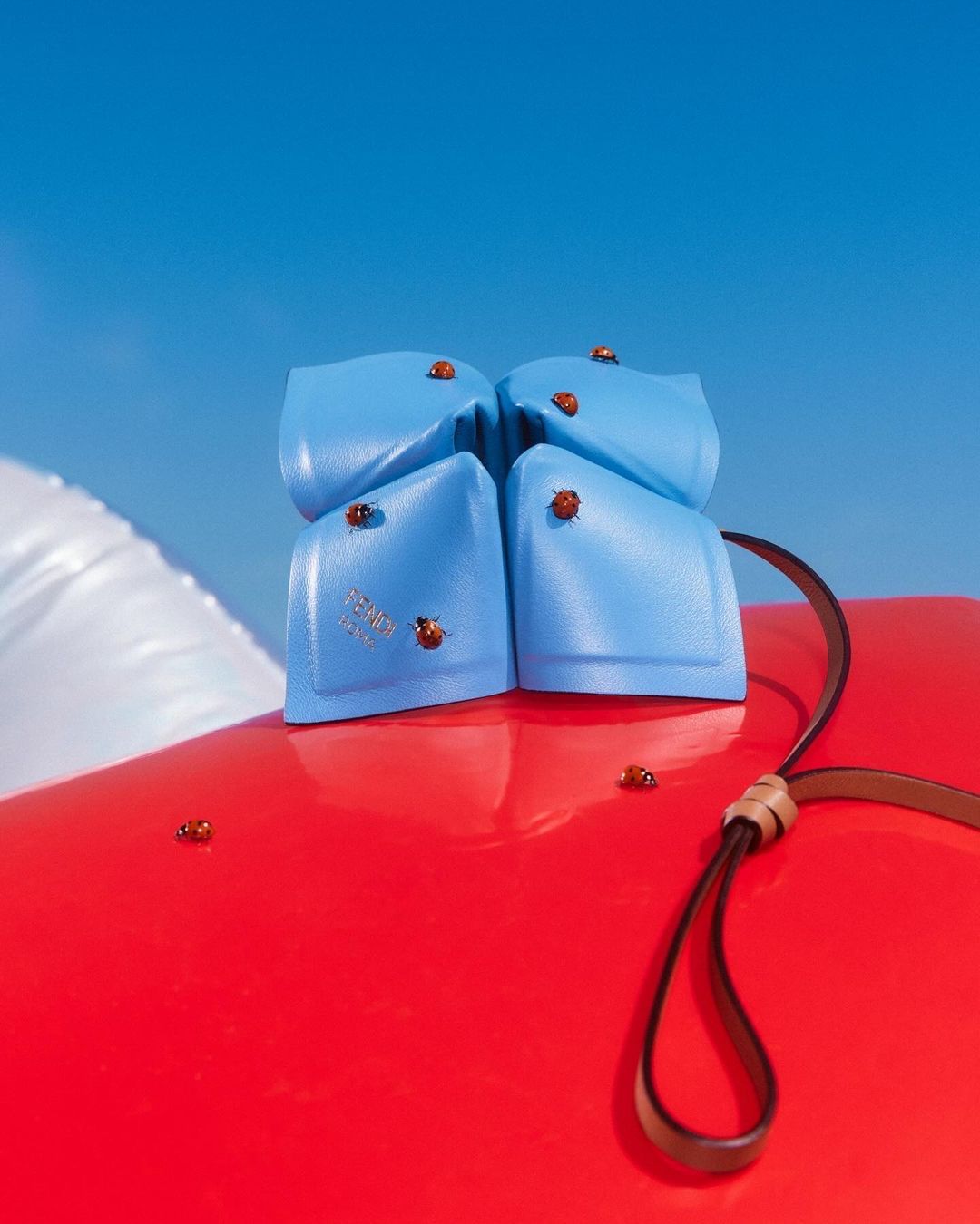 Light blue leather handbag rests on red surface with ladybug accents against a clear blue sky, luxury accessory, product shot.