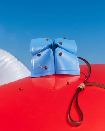 Light blue leather handbag rests on red surface with ladybug accents against a clear blue sky, luxury accessory, product shot.