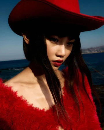 Close-up portrait of a woman wearing a red cowboy hat and a red textured top outdoors against a coastal background.