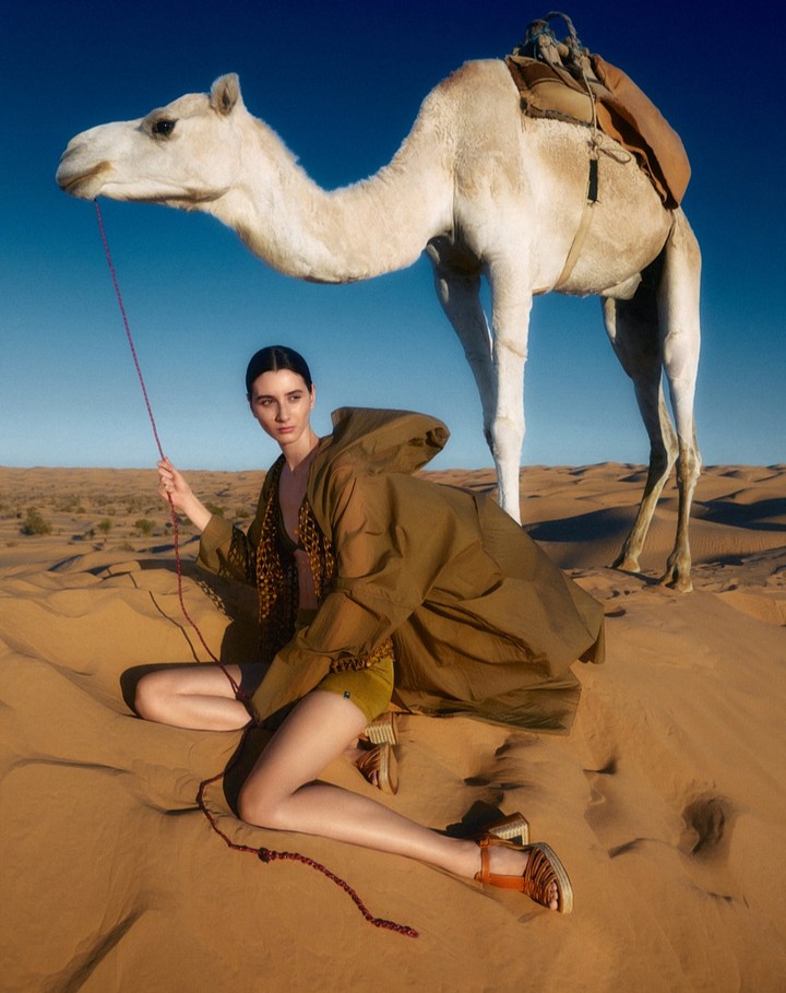 A woman and a dromedary camel in a desert landscape under a clear blue sky. The model holds the camel's lead.