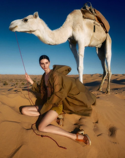 A woman and a dromedary camel in a desert landscape under a clear blue sky. The model holds the camel's lead.