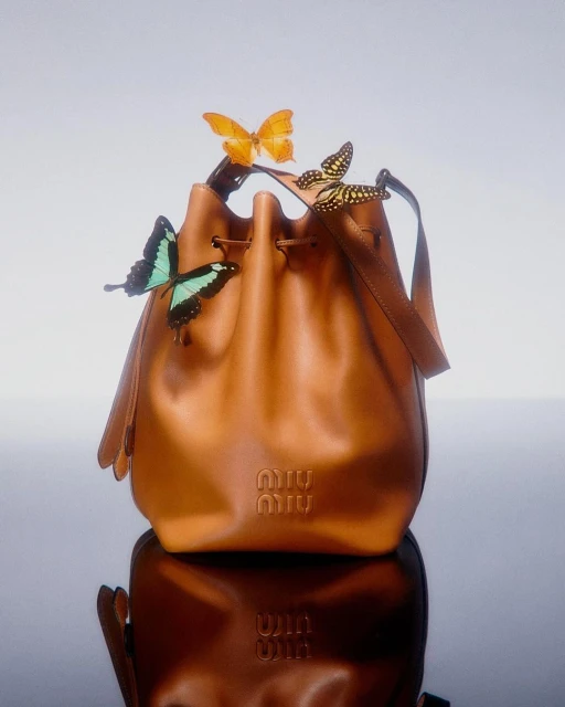 Tan leather Miu Miu bucket bag with three butterflies perched on the top against a soft gradient background and reflective surface.
