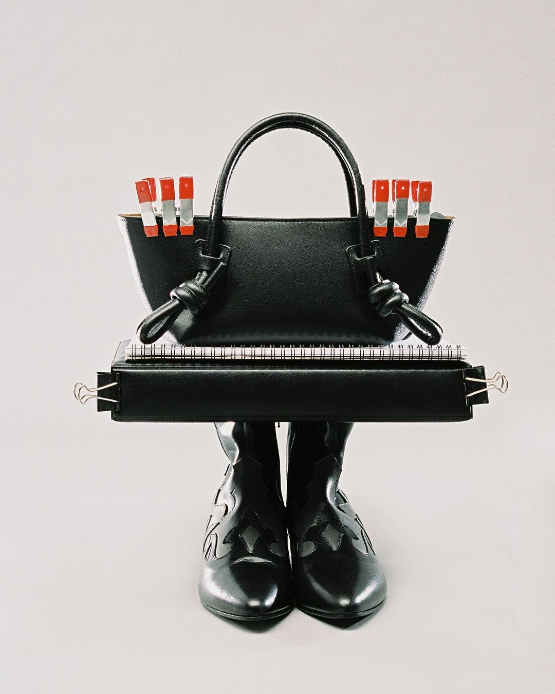 Still life of a black purse with clothespins, notebook, and boots against a plain grey background.