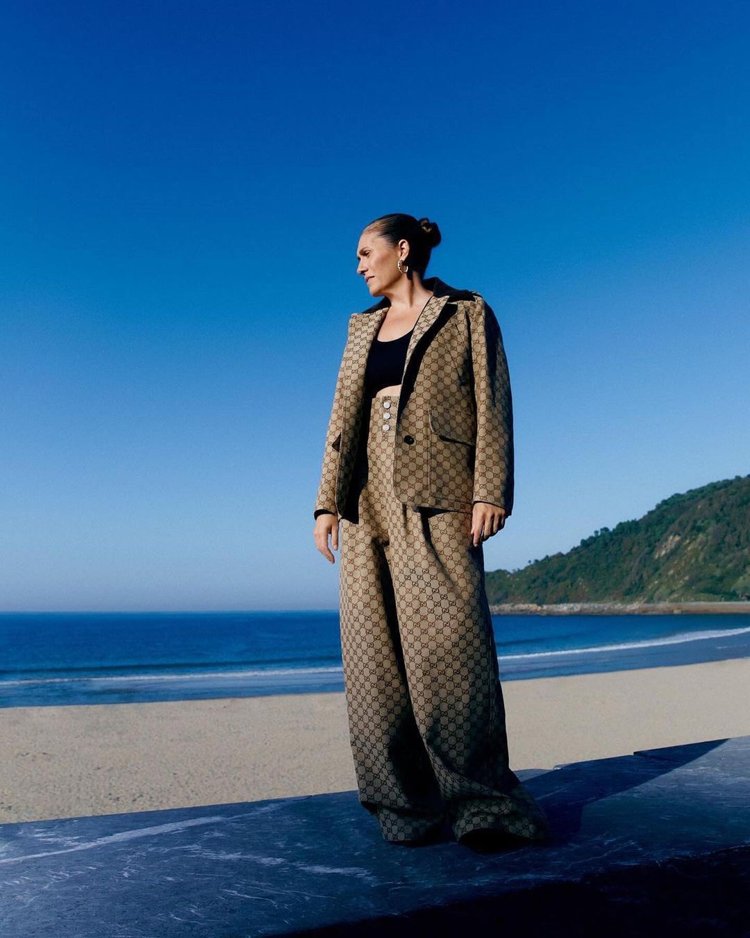 Woman in a brown patterned suit posing on a beach with blue sky and ocean in the background on a bright sunny day outdoors.