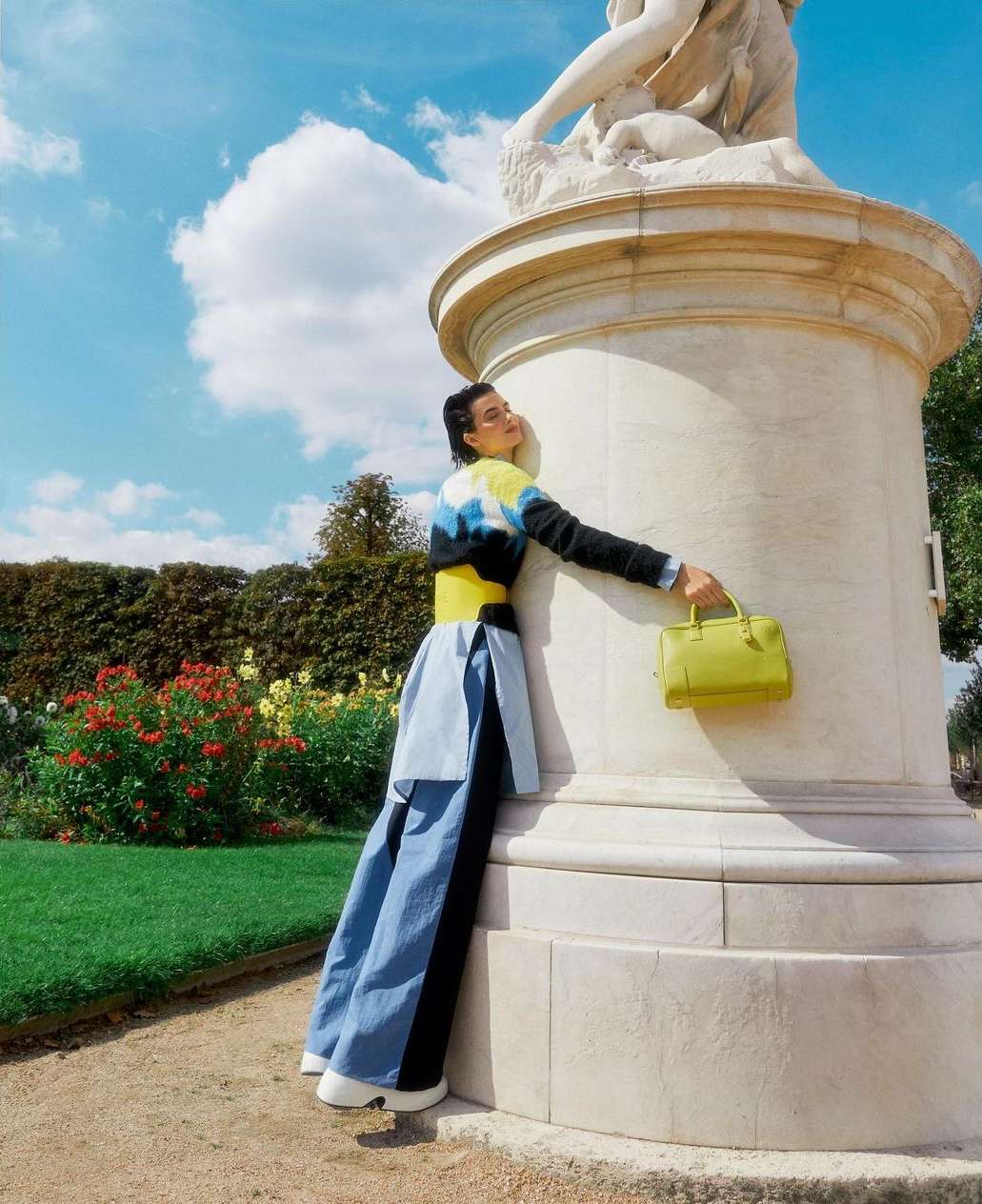 Woman in blue trousers poses next to a stone statue outside. She wears a colourful jumper and holds a lime green handbag.