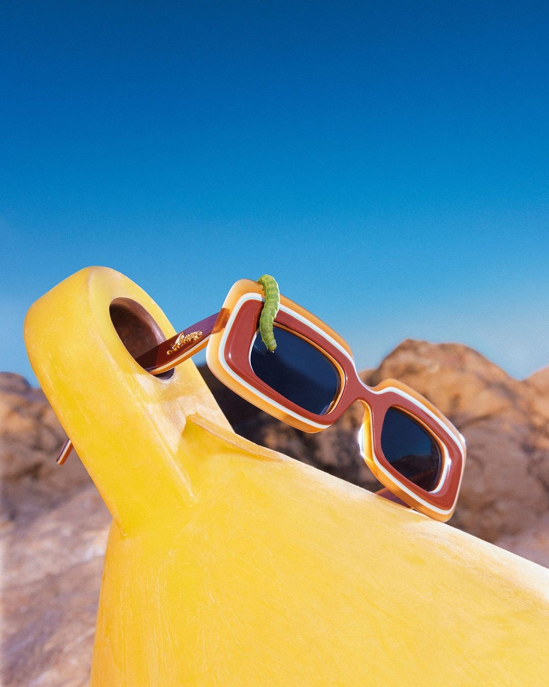 Retro sunglasses perched on a yellow lounge chair under a clear blue sky, with a playful green gummy worm crawling across the frame.