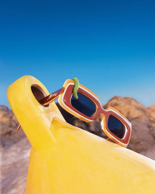 Retro sunglasses perched on a yellow lounge chair under a clear blue sky, with a playful green gummy worm crawling across the frame.