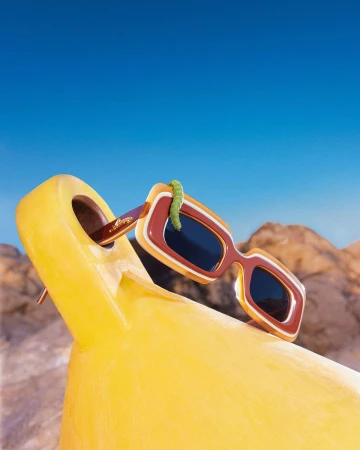 Retro sunglasses perched on a yellow lounge chair under a clear blue sky, with a playful green gummy worm crawling across the frame.