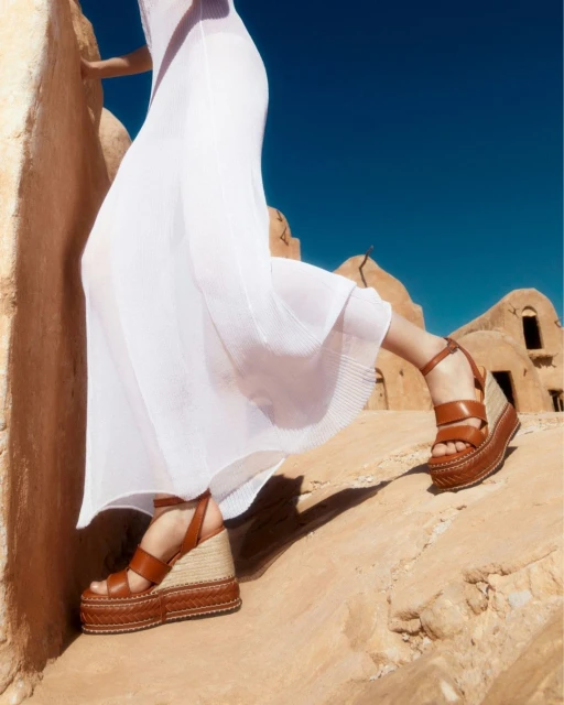 Woman wearing a long white dress and brown leather wedge sandals in an outdoor desert-like setting on a sunny day.