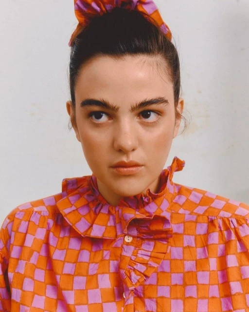Close-up of a young woman with fair skin and dark hair wearing a checkerboard-patterned top and matching hair tie, against a white background.