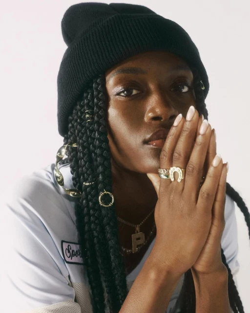 Portrait of a young Black woman wearing a black beanie, statement rings, a gold necklace, and a light blue shirt against a white background.