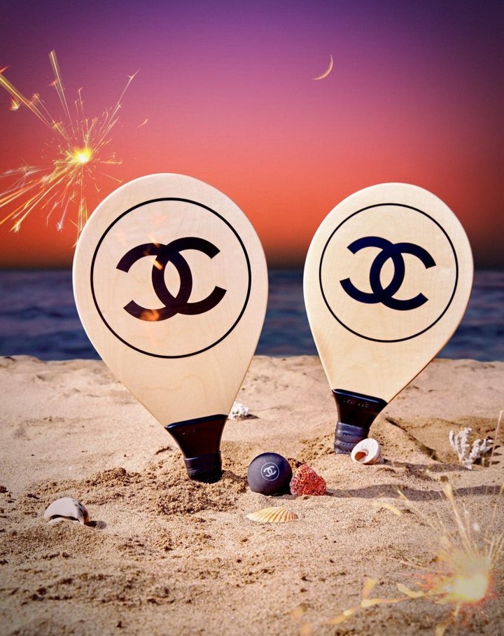 Chanel branded beach rackets with logo on sand at sunset. Beach setting with ocean, pink sky, sparks. Beach and watersports fashion shoot.