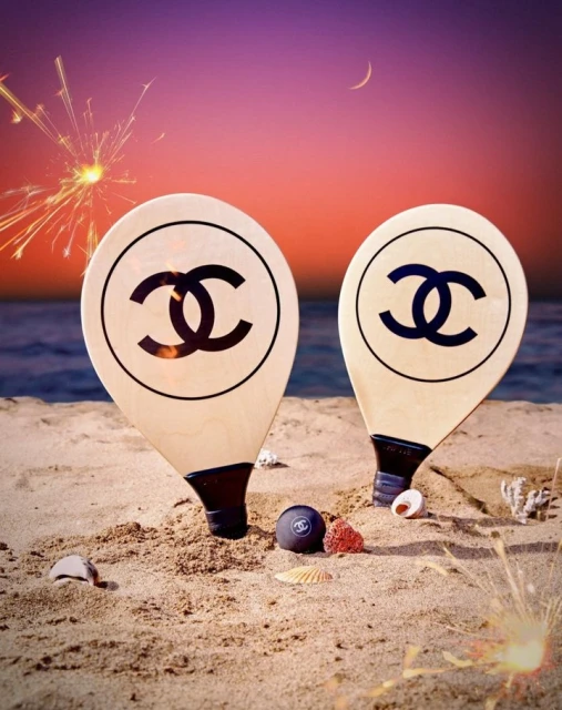 Chanel branded beach rackets with logo on sand at sunset. Beach setting with ocean, pink sky, sparks. Beach and watersports fashion shoot.