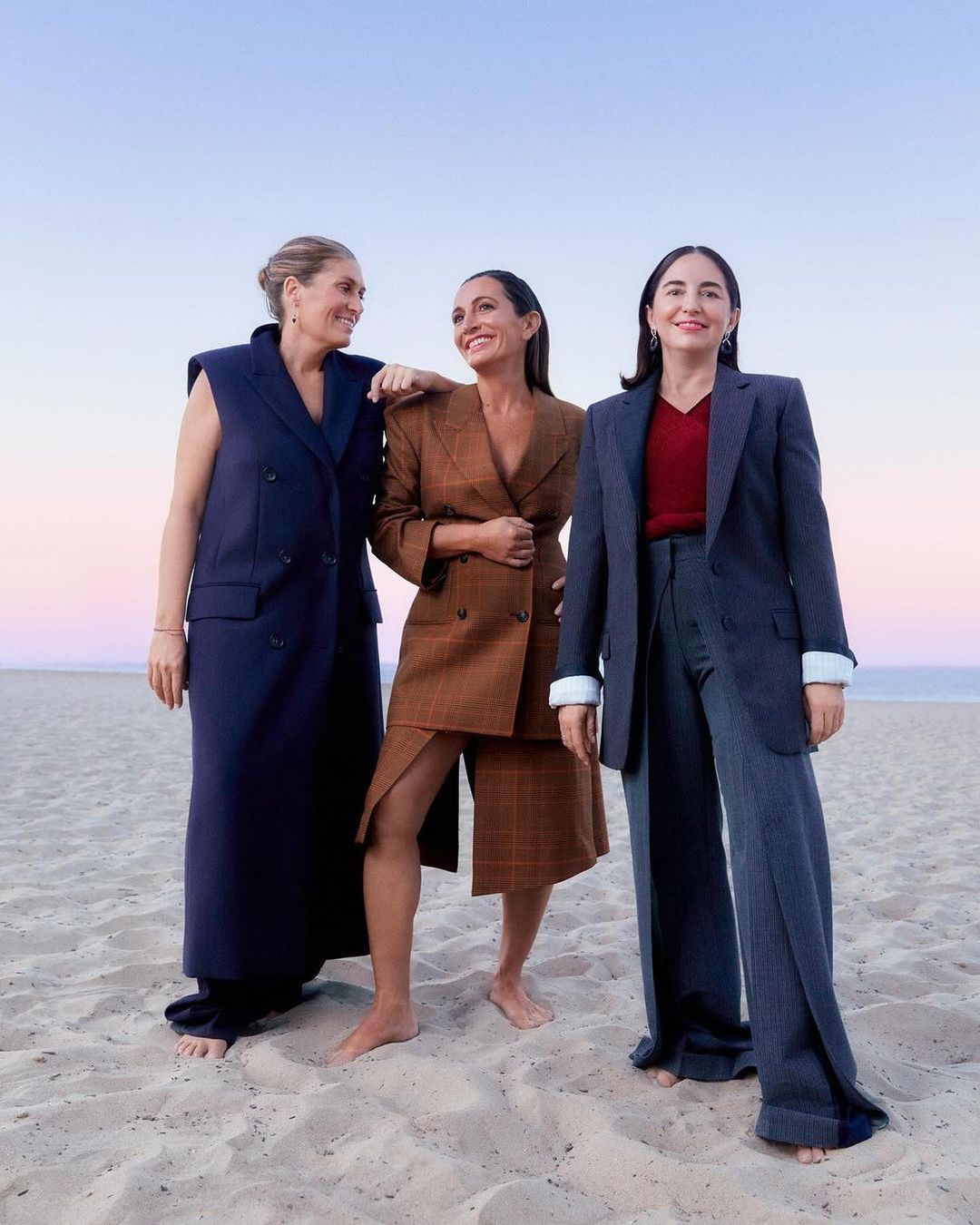Three women in tailored blazers standing barefoot on a sandy beach at sunset by Laia Benavides.