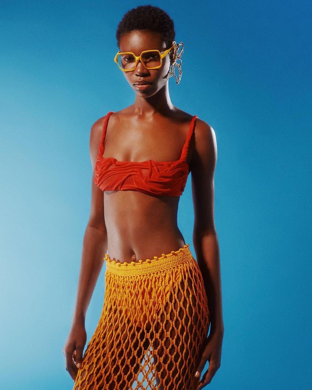 A slender African woman poses confidently against a solid blue backdrop, showcasing a red crop top, yellow net skirt, and bright yellow glasses.
