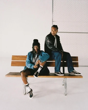 Two models seated on a wooden bench against a white studio background, wearing streetwear and sneakers.