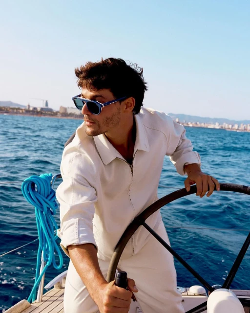Adult male steers a sailboat on the open sea with buildings on the distant shore, wearing sunglasses and a white jumpsuit on a sunny day.