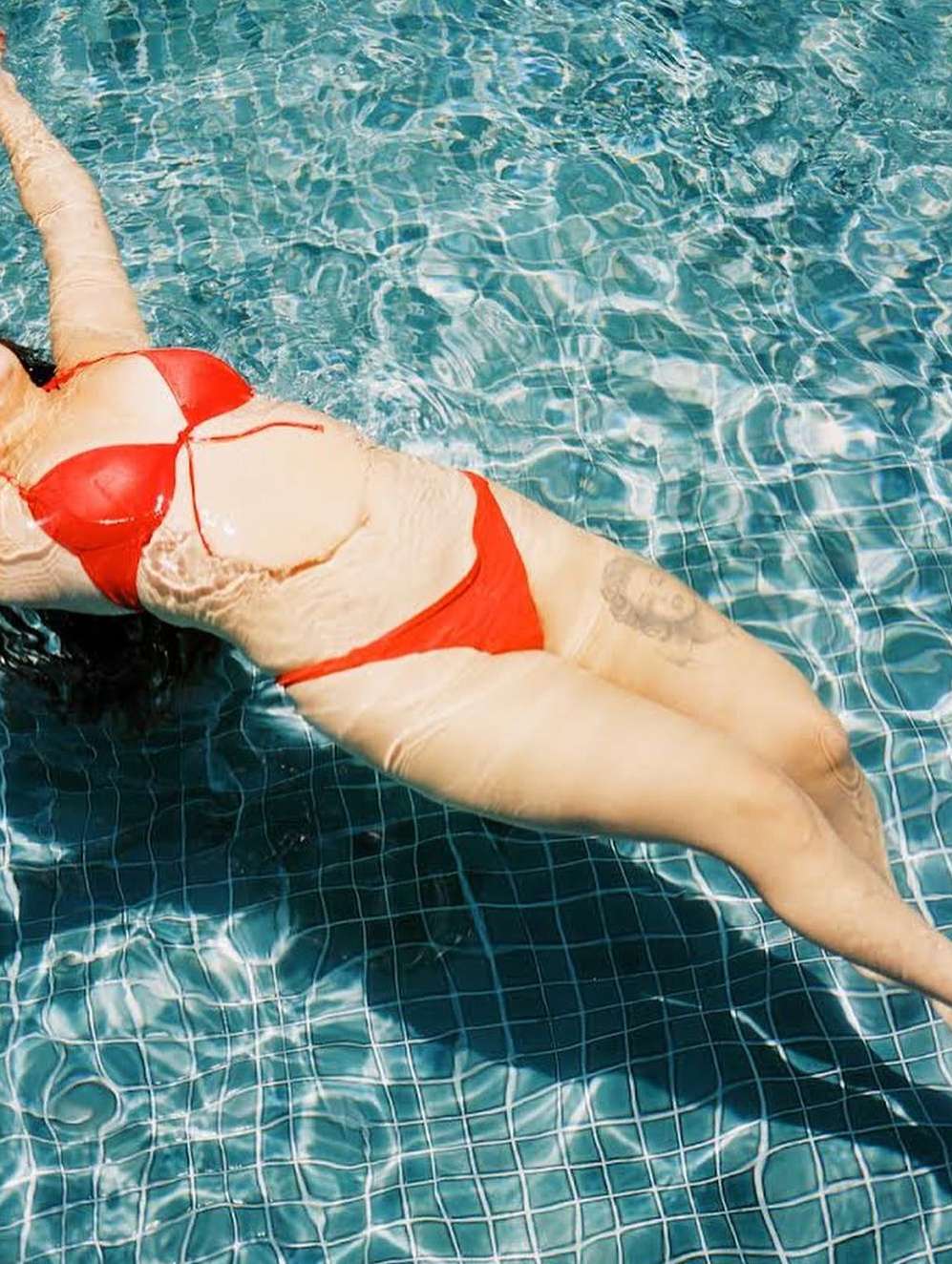 Adult female floats in a pool of blue water with tiled bottom wearing a red two-piece swimsuit. Light reflects off the water.