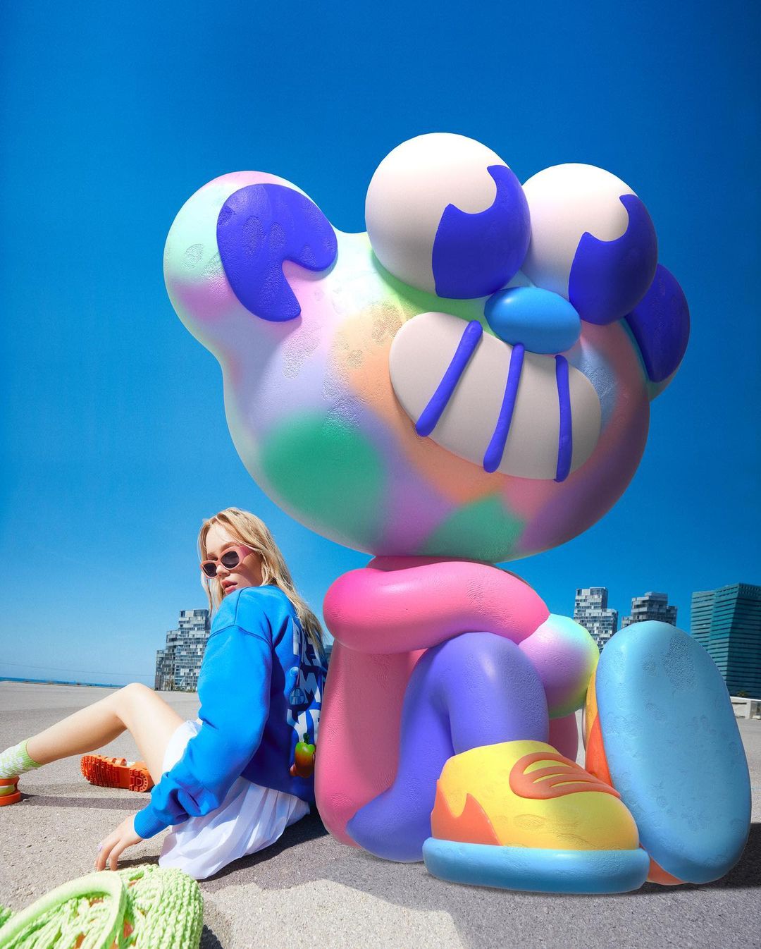 Young woman with sunglasses poses next to an oversized colourful cartoon figure on a city promenade under a bright blue sky.