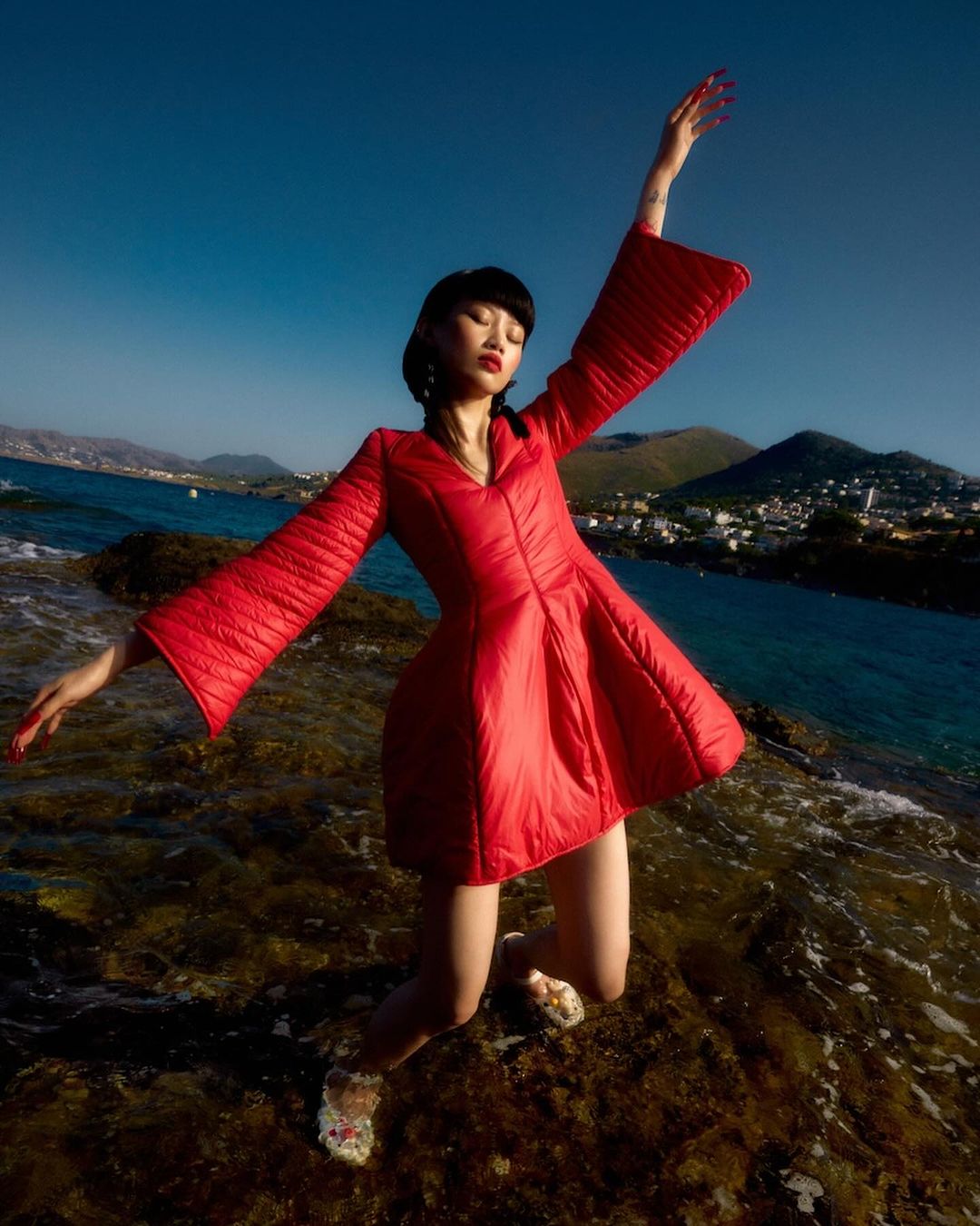 A slender woman poses dramatically in a red dress on a rocky shore with a sea and sky backdrop, illuminated by bright daylight.