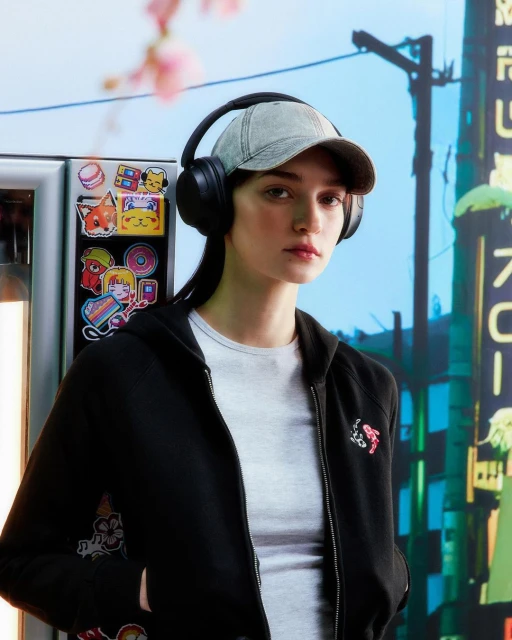 A woman with headphones and a cap poses in front of a vibrant backdrop, wearing a black hoodie and gray shirt. A light adds a glow.