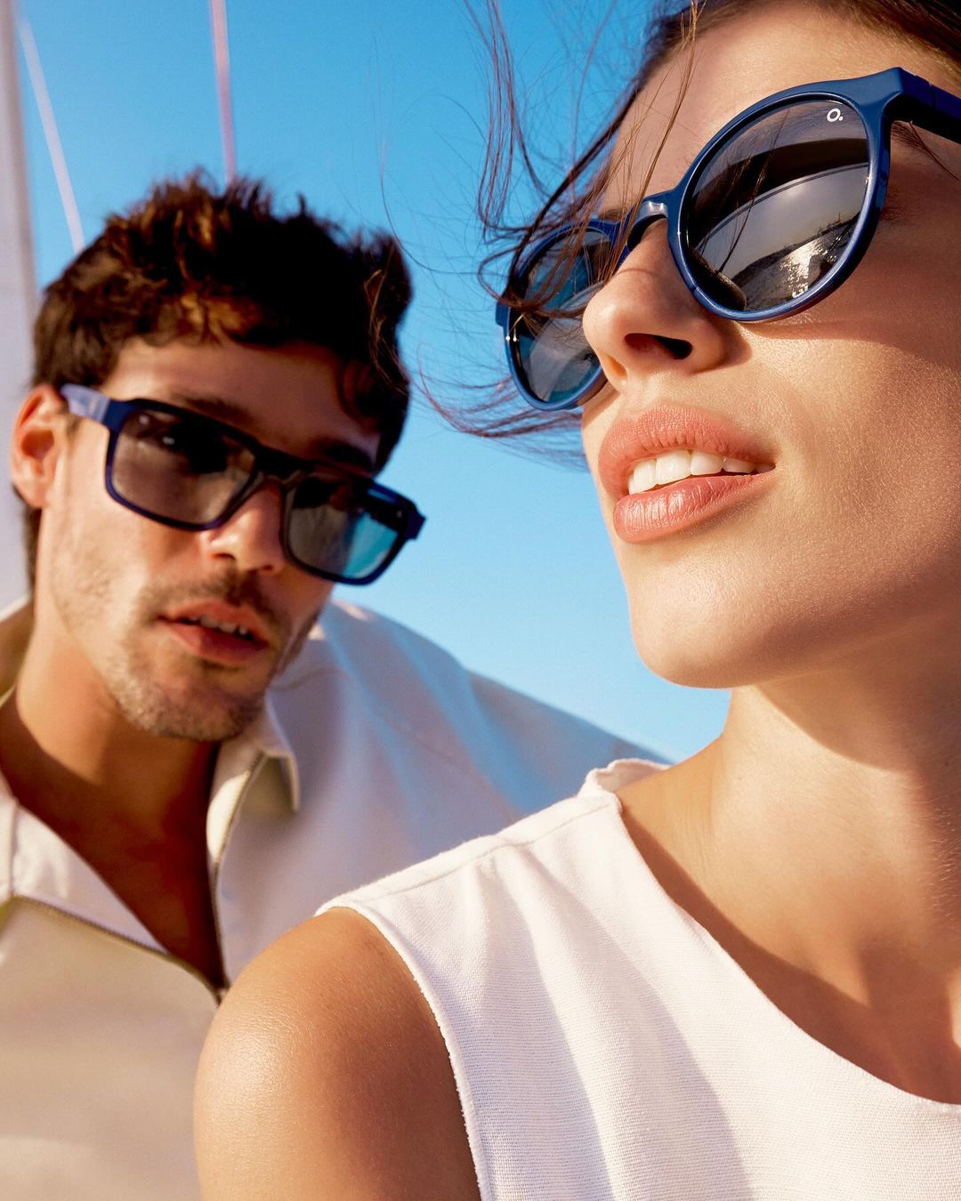 A couple wearing sunglasses is on a sailboat under a bright blue sky. The woman has round blue frames, and the man has rectangular sunglasses.
