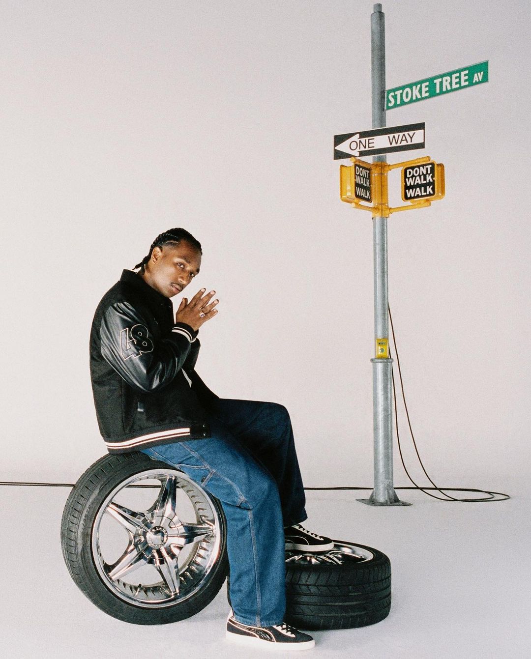 Adult male in a black varsity jacket and blue jeans poses seated on a chrome tire with a street sign prop in a studio setting.