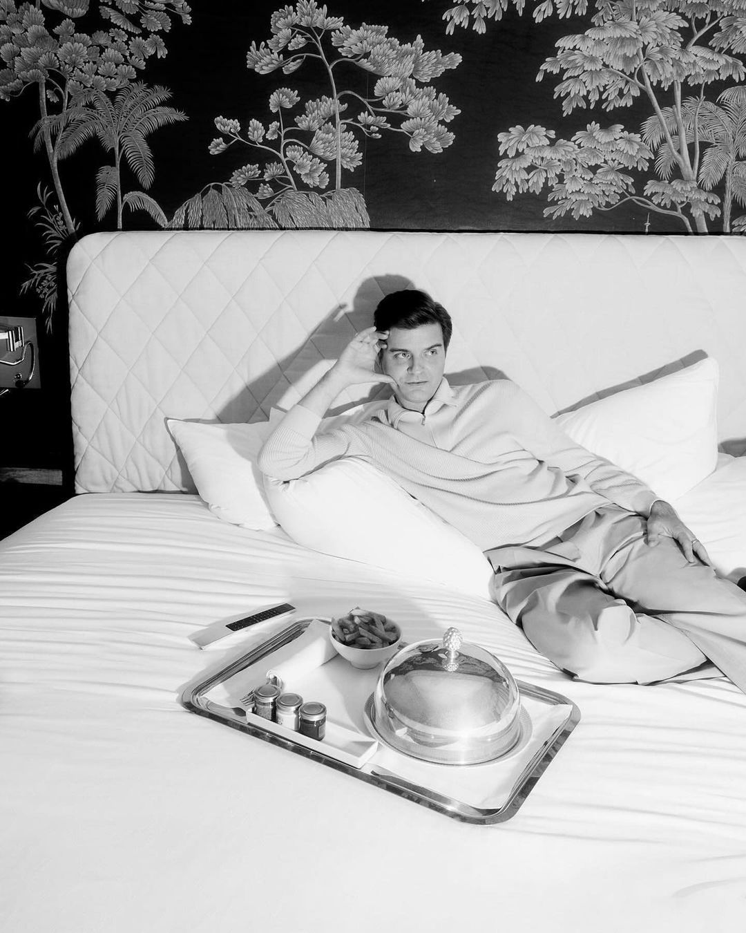 A man in a sweater is lying in bed with a tray of food, resting his head on his hand, with patterned wallpaper behind him.