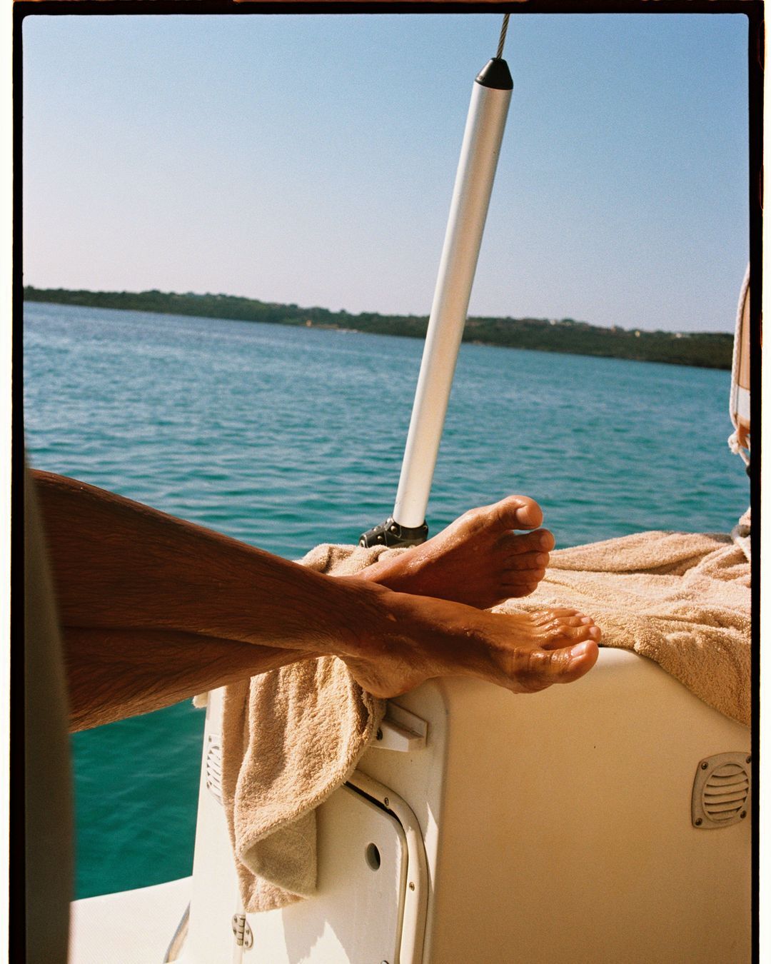 Bare feet rest on the back of a boat, sunny day, with teal water and distant shoreline visible in background under a clear, bright sky.