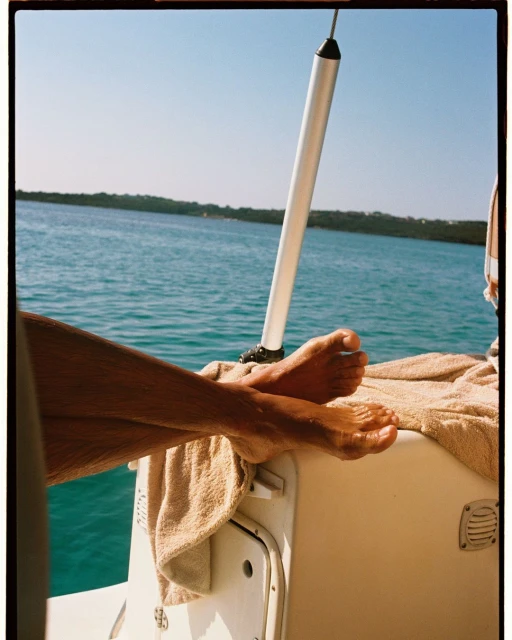 Bare feet rest on the back of a boat, sunny day, with teal water and distant shoreline visible in background under a clear, bright sky.
