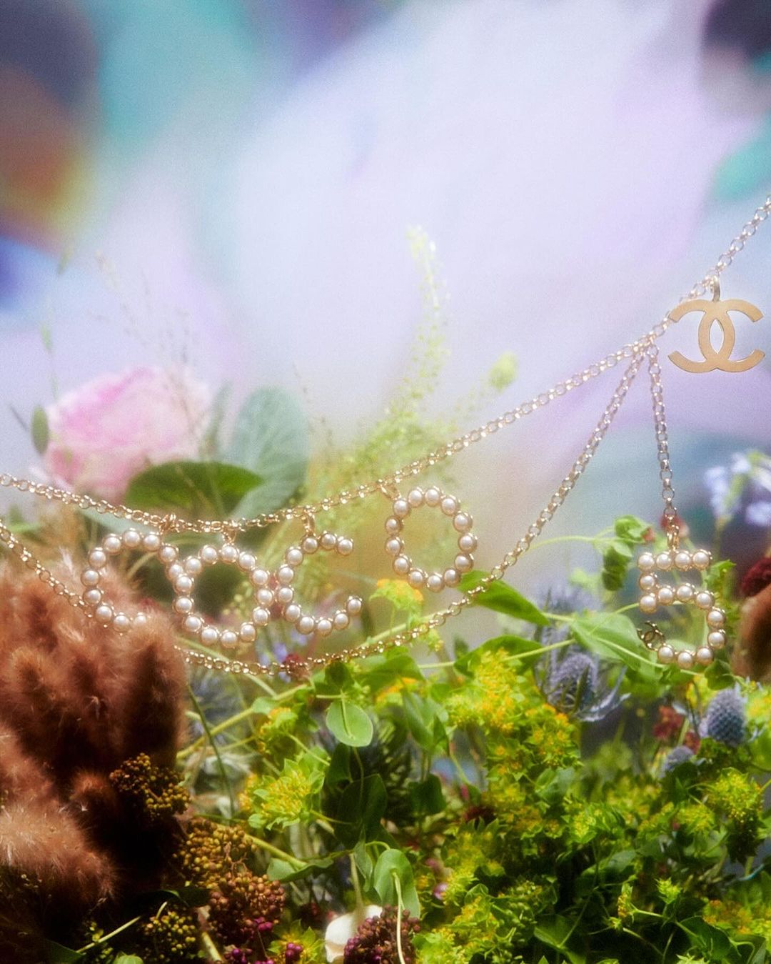 Close-up of a gold Chanel necklace spelling 'COCO' with pearls over green foliage and blurred flowers in a soft focus, ethereal style.