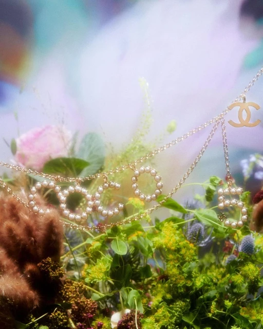 Close-up of a gold Chanel necklace spelling 'COCO' with pearls over green foliage and blurred flowers in a soft focus, ethereal style.