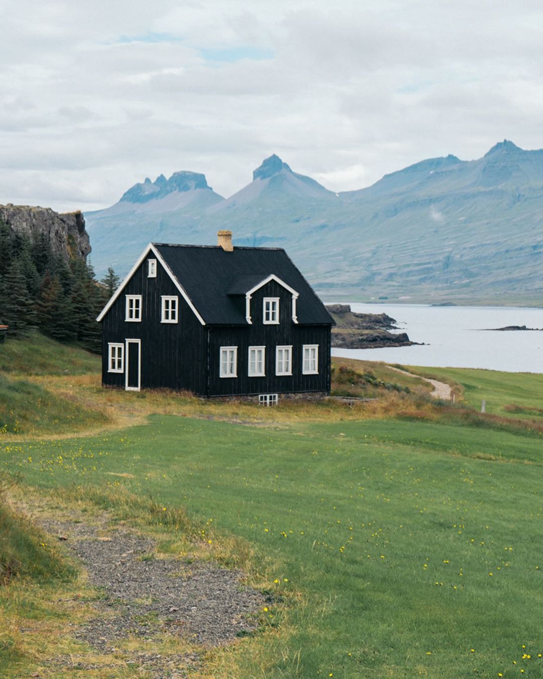 Scenic view of a black wooden house in a green field with mountains and a body of water in the background under an overcast sky.