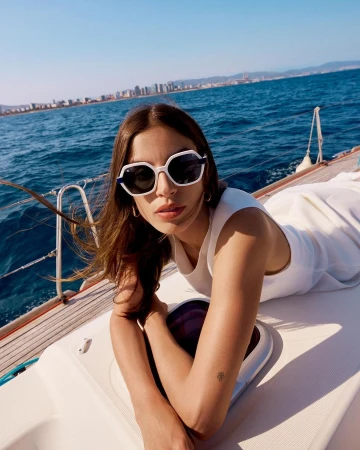 A woman wearing white sunglasses and a white dress lying on a sailboat deck with the ocean in the background